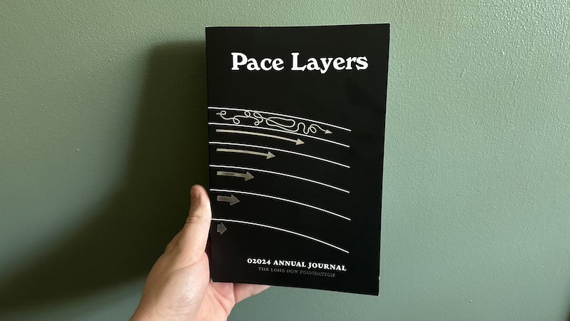 A person holds up a copy of Pace Layers against a green wall.